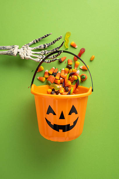 istock Orange pumpkin bucket overflowing with assorted Halloween candy on vertical green backdrop. Skeleton hand adds a spooky touch, embodying Halloween festivity 2229995146