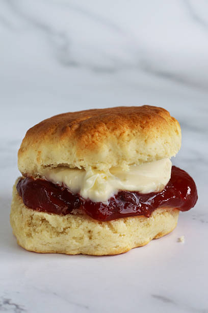 istock Image of individual plain scone with Cornish clotted cream and strawberry jam, traditional English afternoon tea, baked good topped with jam and cream the Cornish way, white marble effect background, focus on foreground 2229995711