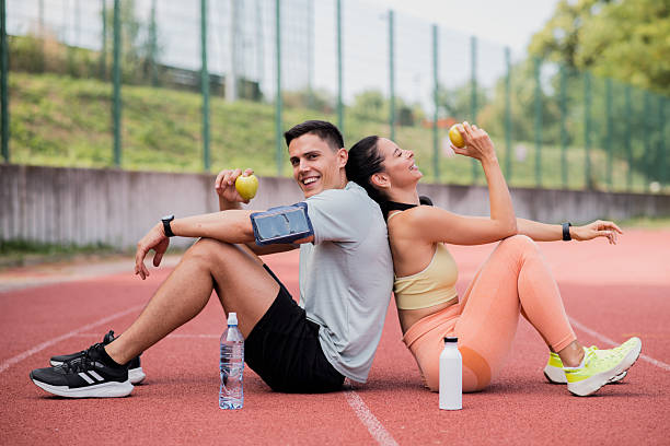 Muscular young man and beautiful young woman sitting on an outdoor track back to back, laughing and eating apples together after a run.