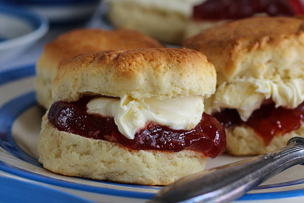 istock Full frame image of plain scones with strawberry jam and clotted cream on blue and white striped Cornish ware plate, knife, traditional English afternoon tea topped with jam and cream the Cornish way, focus on foreground 2229999798