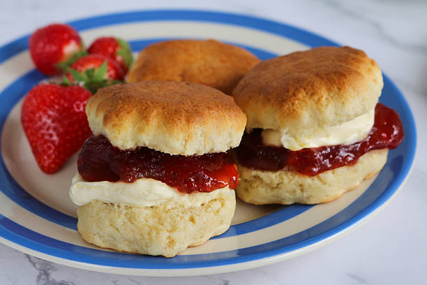 istock Close-up image of plain scones with strawberry jam and clotted cream on blue and white striped Cornish ware plate, strawberries, traditional English afternoon tea topped with jam and cream in the Cornish and Devonshire ways, social debate concept 2229999800