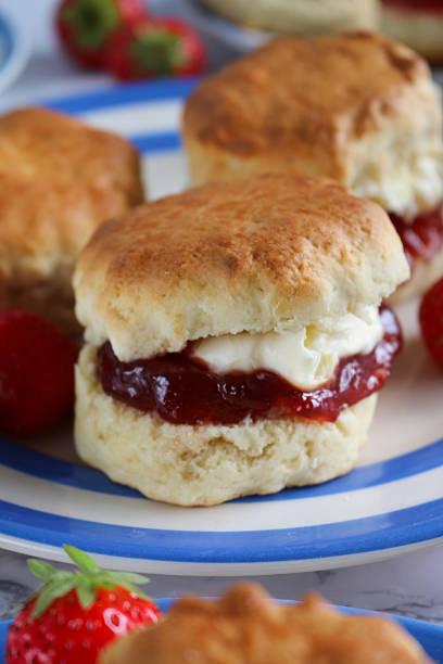 istock Close-up image of plain scones with strawberry jam and clotted cream on blue and white striped Cornish ware plate, strawberries, traditional English afternoon tea topped with jam and cream in the Cornish and Devonshire ways, social debate concept 2229999914