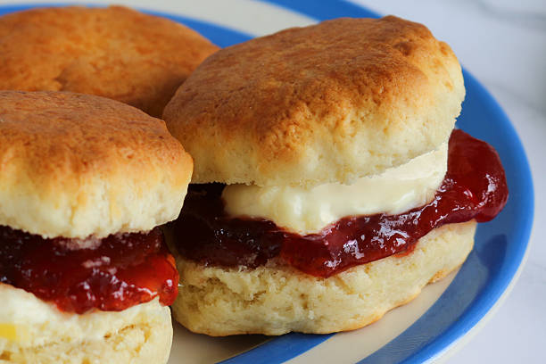 Stock photo showing close-up view of an English afternoon tea scene, featuring a blue and white striped Cornish ware plate containing golden, plain scones filled with clotted cream and strawberry jam. The scones are filled in both the Cornish way with jam spread on the scone first, followed by a dollop of clotted cream, and the Devonshire way with cream first with a spoonful of jam on top. Social debate concept.
