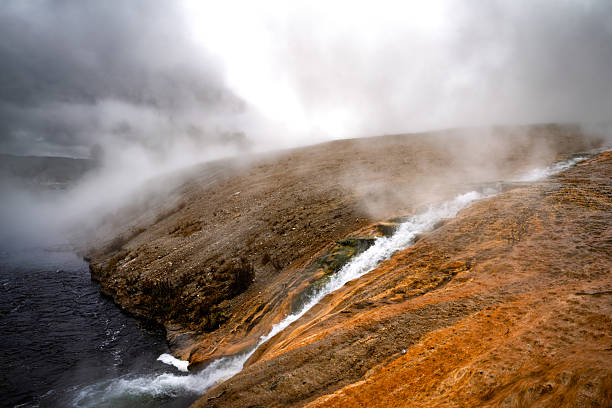 Scalding thermal water flows from a geothermal spring down a rocky slope and into a river under heavy steam in Yellowstone National Park. The vivid orange and rust-colored mineral deposits contrast with the dark water and cloudy sky, capturing the park’s otherworldly and volatile geothermal landscape.