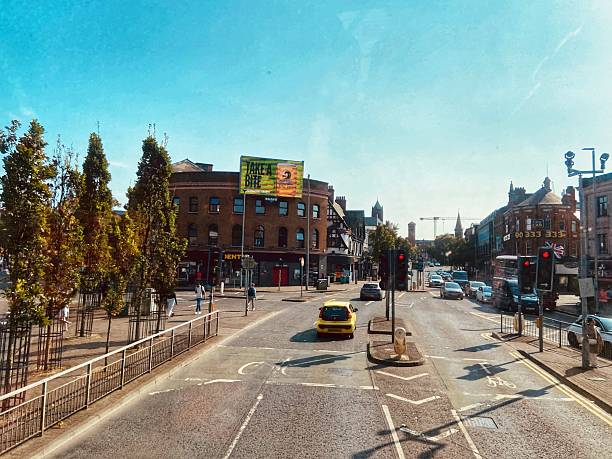 Belfast, United Kingdom - August 12, 2025. A busy urban intersection with a yellow car stopped at a red traffic light. Brick buildings, trees, and a large advertisement are visible.