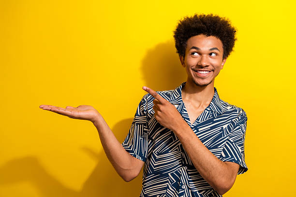 istock Smiling young man in casual patterned shirt pointing and presenting gesturing cheerfully against a vibrant yellow background 2230064419