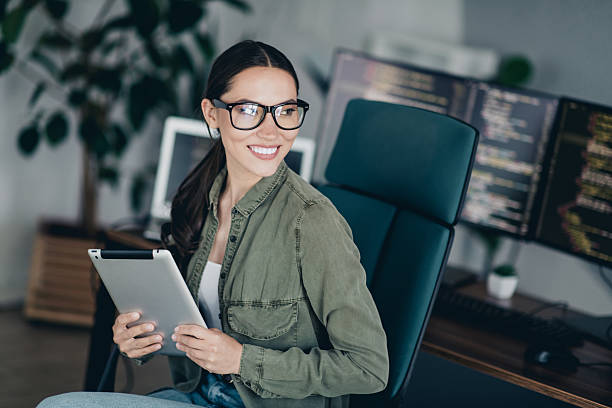 istock Young female programmer working in a modern office, smiling confidently while holding a tablet, surrounded by computers 2230064578