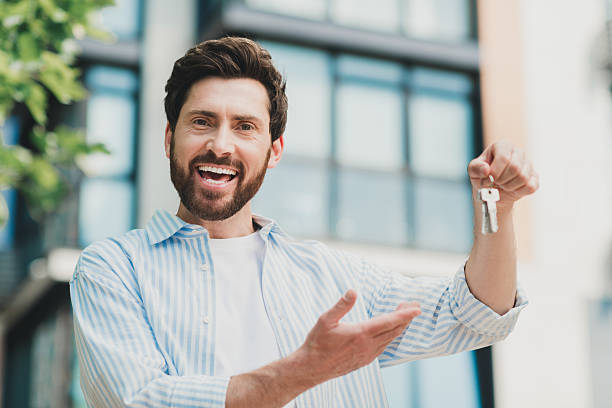 istock Young man holding keys outdoors, smiling confidently while gesturing towards buildings in a sunny urban cityscape setting 2230066137