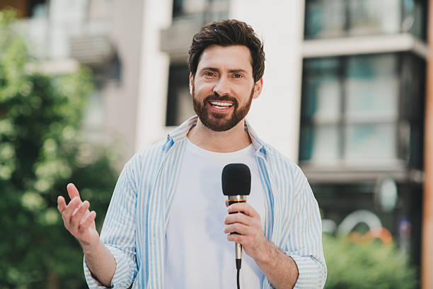 istock Smiling young man presenting with a microphone outdoors beside urban buildings and greenery on a sunny day 2230066145