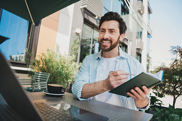 istock Young man outdoors taking notes in a journal, smiling and surrounded by greenery and sunlight at a street café 2230066197