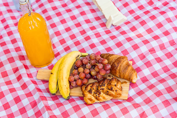 Freshly baked croissants, ripe bananas, and juicy grapes arranged on a wooden board with a refreshing bottle of orange juice create a delightful picnic spread on a red and white checkered blanket