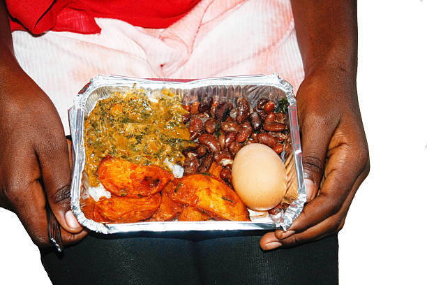 Close-up of a traditional Rwandan meal served in a foil takeaway container, featuring fried plantains, boiled egg, beans, and vegetable sauce over rice, held in a person’s hands. Captured in Rwanda, showcasing authentic African cuisine and local food culture.