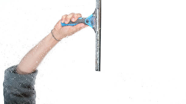 A Caucasian female hand holds a blue and black window squeegee wiping water from a glass panel, used for cleaning windows or transparent partitions.