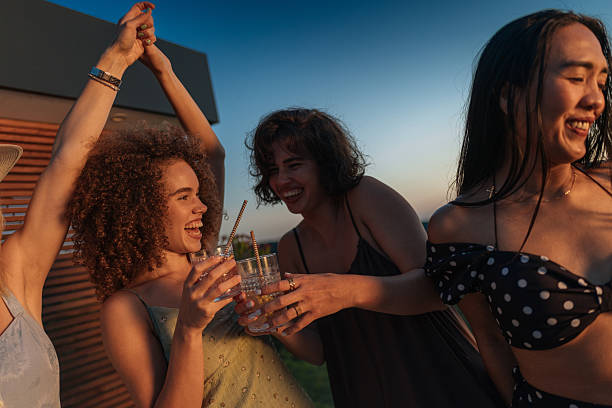 Group of young women laughing and having fun together at a rooftop party, raising their glasses for a toast during sunset