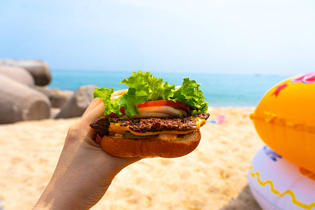 istock Woman holding burger in a hand against beach background 2230111054