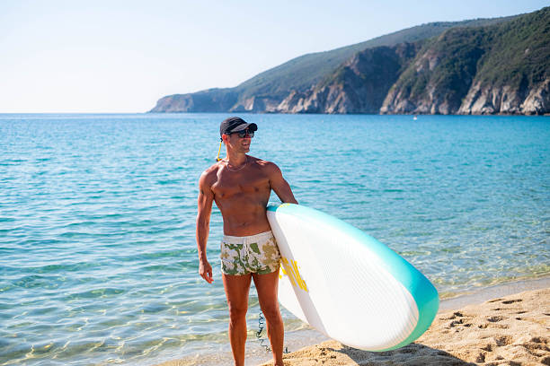 muscular man holding a surfboard on the beach on the seashore, in a suit, wearing sunglasses, he is happy