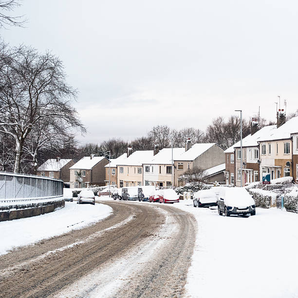 Deep snow covering the parked vehicles and houses on a street curving down a hill.
