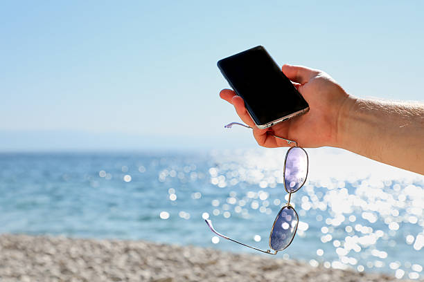 Smartphone and sunglasses held by a human hand against tranquil ocean and bright sky, perfect for leisure, travel, lifestyle, and mobile connectivity themes.