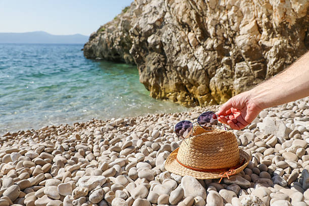 Straw hat with sunglasses placed on a pebble beach by the turquoise sea and rocky cliffs, while a male hand gently adjusts the glasses, symbolizing summer relaxation.