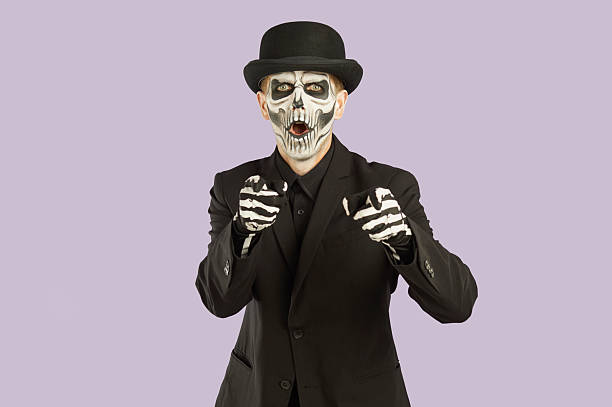Amazed man with skeleton face pointing two forefingers at camera. Portrait of handsome man with scary Halloween make up wearing bowler hat and black suit pointing forward on isolated studio background