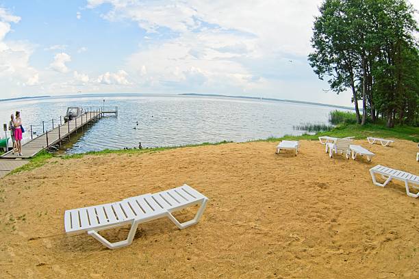 Lake Narach, Belarus, July 16, 2025.  Sandy beach with several white plastic lounge chairs scattered on the shore near a wooden pier extending into the water. A person is standing on the pier, gazing at the lake, which has a calm surface. The landscape is bordered by trees on the right, and the sky is partly cloudy. The wide-angle lens distorts the perspective, creating a sweeping view of the scene.