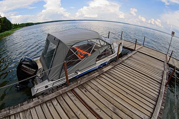 Lake Narach, Belarus, July 16, 2025.A motorboat is docked at a wooden pier on a serene lake. The boat has a gray canopy and clear side windows, with an outboard Mercury motor. Orange life jackets are visible inside. The wooden planks of the pier are weathered, stretching out over calm water. Tall reeds are visible to the left, and a dense line of trees borders the lake. Fluffy clouds scatter across a partly sunny sky, reflecting on the lake s surface, suggesting a peaceful, outdoor scene.