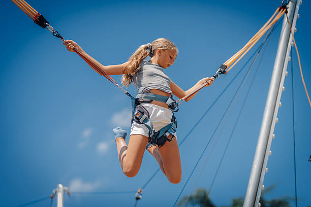 Girl wearing safety harness enjoying bungee jumping trampoline against blue sky in amusement park