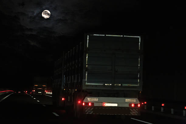 Car point of view looking through windshield at a solitary late night expressway speeding semi tractor trailer truck hauling container freight on an otherwise deserted midnight highway. The car is entering the multiple lane highway to merge from the right after a curving entrance ramp road.