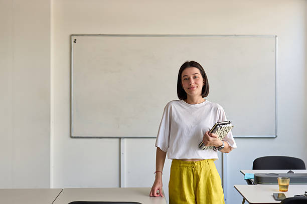 istock Young teacher standing in classroom holding notebooks 2230166819