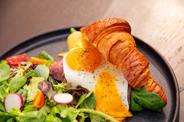 Gourmet brunch plate from an Istanbul cafe featuring a buttery croissant filled with sunny side up egg, melted cheddar cheese, smoked meat and fresh greens. Served with radish, cherry tomatoes and colorful salad. Ideal for breakfast menus, café promotions and healthy lifestyle content.