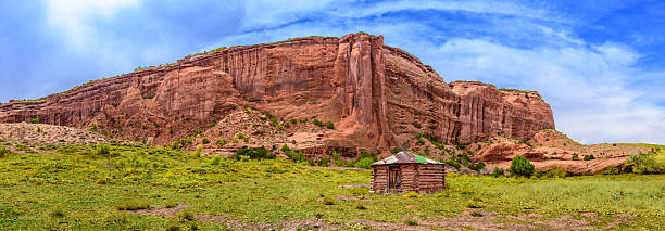 The hogan is the traditional dwelling of the Navajo people, designed to harmonize with the surrounding landscape and cultural beliefs. Built from logs, wooden poles, and packed earth, with a door always facing east to greet the rising sun, the hogan serves not just as shelter but as a spiritual space. There are several styles, but the type seen here is a simple wooden variant, often used as a home or ceremonial structure. This traditional Navajo hogan stands beneath the towering sandstone cliffs of Canyon de Chelly, a reminder that this sacred canyon is still home to Navajo families who maintain a deep connection to their ancestral land. Many Navajo families within Canyon de Chelly still live in or maintain hogans, blending traditional life with modern influences. The canyon itself has been continuously inhabited for thousands of years, making it one of the longest continuously occupied landscapes in North America.  This hogan was photographed by the Canyon Road in Canyon de Chelly National Monument near Chinle, Arizona, USA.
