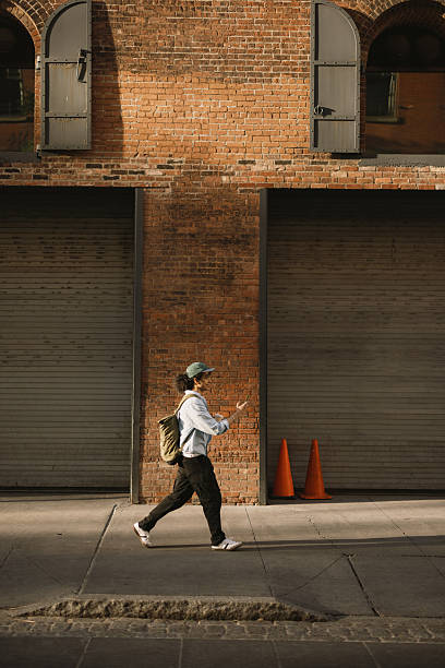 Young man walking on the street and using his phone in front of a brick wall building in New York