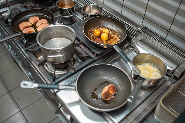 A professional kitchen showcasing a stainless steel stove with multiple pans and pots cooking meat, eggs, pasta, and sauces. The organized layout and active cooking scene emphasize a busy culinary setting.