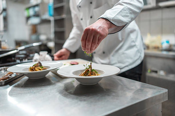 A male chef in a white double-breasted jacket adds fresh garnish to a plated dish in a sleek, modern kitchen, showcasing culinary artistry and precision in a professional environment.