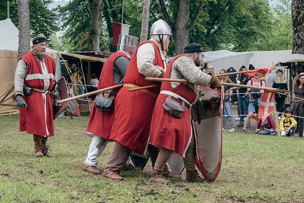 Tuttlingen, Baden-Wuerttemberg, Germany – 6.10.2022: Four people dressed in medieval-style attire participate in an outdoor historical reenactment. They wear red robes and wield large shields and weapons, appearing as if in formation. The scene is set in a grassy area with trees, tents, and spectators in the background. The event is lively, with onlookers gathered around to watch. The attire, featuring helmets and robes, evokes the medieval era, emphasizing a historical or cultural festival atmosphere.