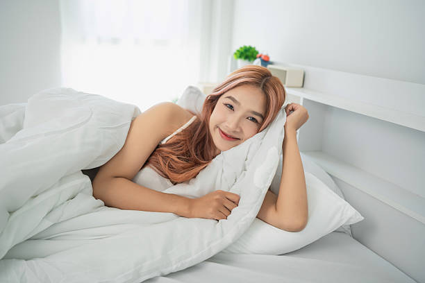 istock Young Woman Relaxing in Bed with Soft White Bedding, Enjoying Morning Light, Smiling and Pampering Herself in Cozy Bedroom Environment 2230210010