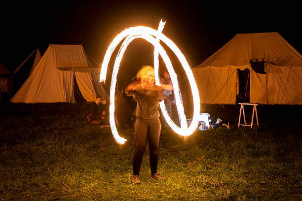 Nordrach, Baden-Wuerttemberg, Germany – 4.8.2023: A woman performs a fire show at night, creating swirling patterns of light. She stands on a grassy area in front of several large, beige canvas tents. The setting suggests an outdoor camping or festival event. The tents and the woman's attire are illuminated by the fire's glow, highlighting her motion against the dark night backdrop. A few other people are visible, sitting and observing the performance.