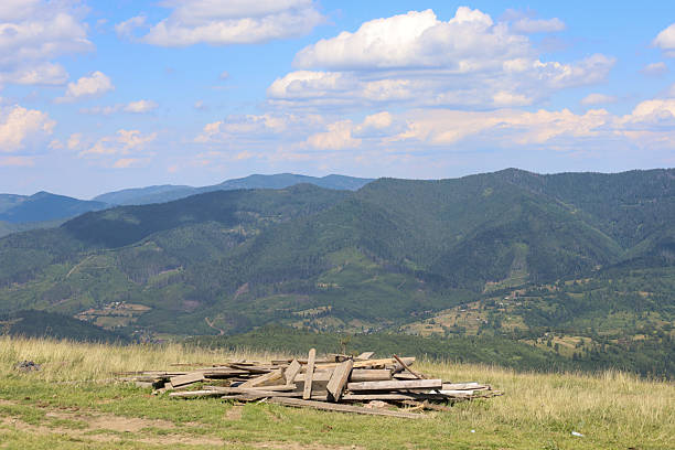 ​A breathtaking mountain landscape unfolds under a clear blue sky with fluffy white clouds, showcasing a lush, green valley and a distant forest-covered range. In the foreground, a pile of rough-hewn wooden planks rests on a sun-drenched grassy hill.