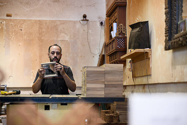 Middle Eastern craftsman holding up a wooden frame for close inspection inside a rustic studio, with shelves, tools, and traditional decor in the background.