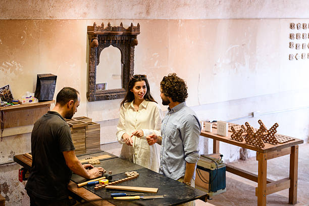 Candid shot of visitors engaged in conversation with a craftsman inside an Arabic wood workshop, standing by a bench with chisels, timber panels, and decorative wooden pieces.