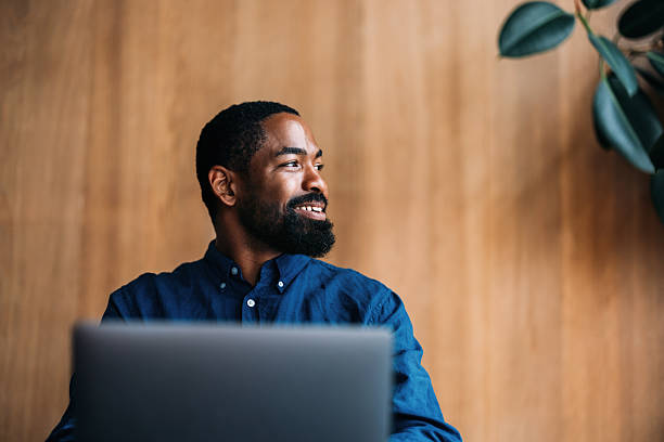 A confident professional man sits working on his laptop in a modern office space, exuding positivity and reflecting a workplace atmosphere. The background is minimalist with a plant adding natural decor.