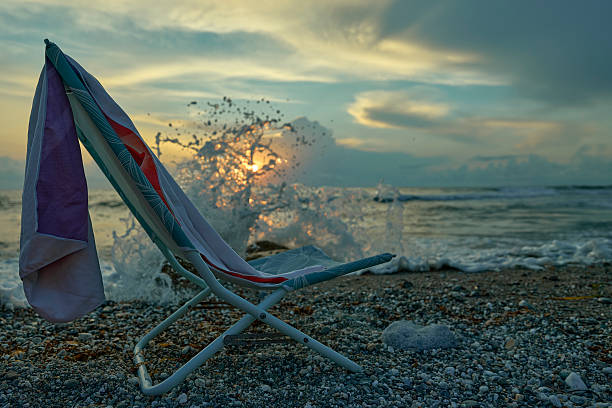 A beach chair with a towel facing a sunset over the ocean with waves splashing on the shore.