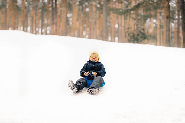 Smiling child enjoying sledding downhill on a snowy slope in a winter forest, having fun during winter holidays