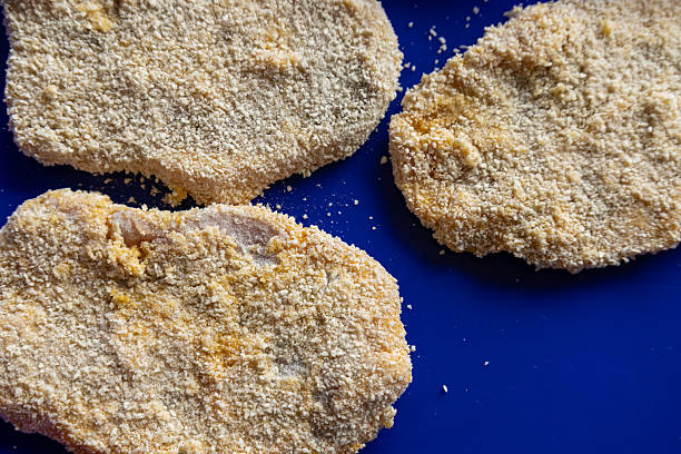 Top view of three raw breaded pork loin slices arranged on a dark blue tray. Homemade preparation of breaded fried pork loin.