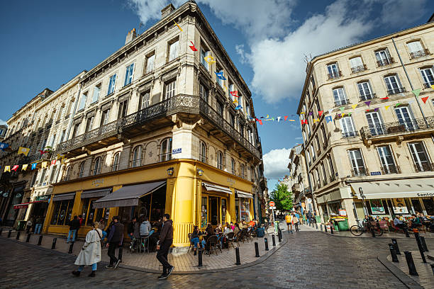 Bordeaux city street showcasing historic buildings under a vibrant sky. A bustling café accentuates the lively ambiance, adorned with colorful flags and visitors enjoying an outdoor urban setting.