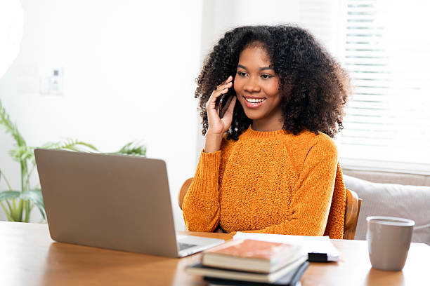 istock A smiling African-American woman is seen talking on her mobile phone while using a laptop. She is sitting at a desk with a mug and notebooks nearby 2230274009