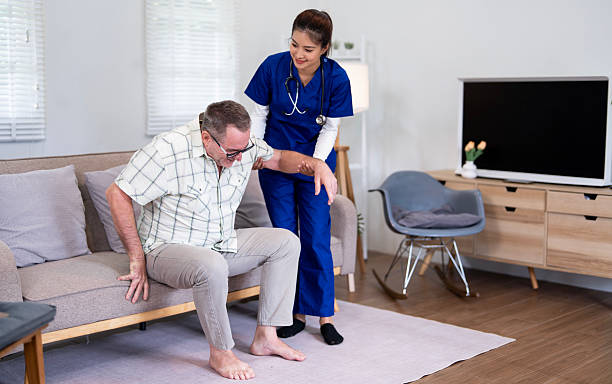 istock Nurse in blue scrubs assisting elderly man to stand up from sofa, providing mobility support and home healthcare assistance. 2230274558