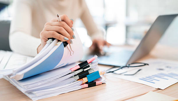 istock A woman is seen multitasking, holding a stack of organized documents with a pen while working on a laptop. 2230275164