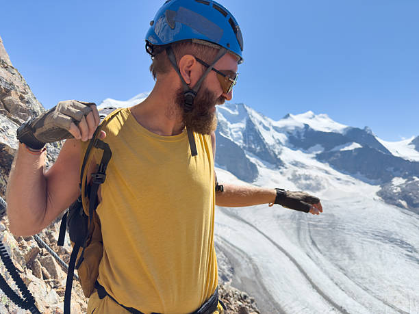 Bearded man wearing helmet and harness climbing a steep rocky mountain with snowy peaks and glacier in the background. Outdoor adventure and extreme sports enthusiast on mountain climb. Mountain climbing activity in alpine environment.