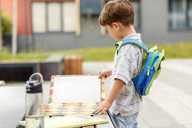 Young boy stands outdoors near bench, examining colorful school books with water bottle beside him on a sunny day.